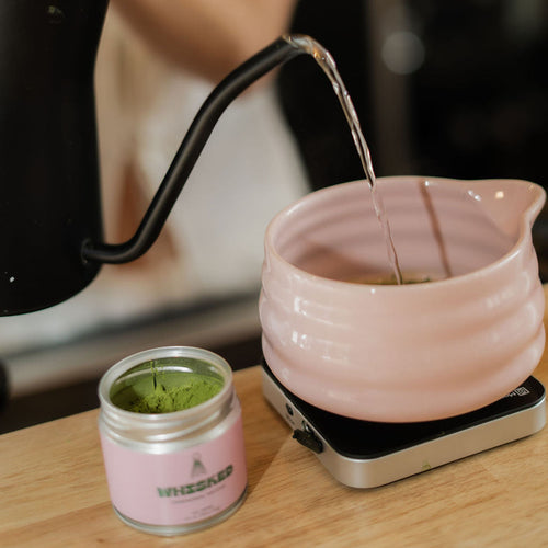 Person pouring water from a black kettle into a pink teapot on a wooden table.